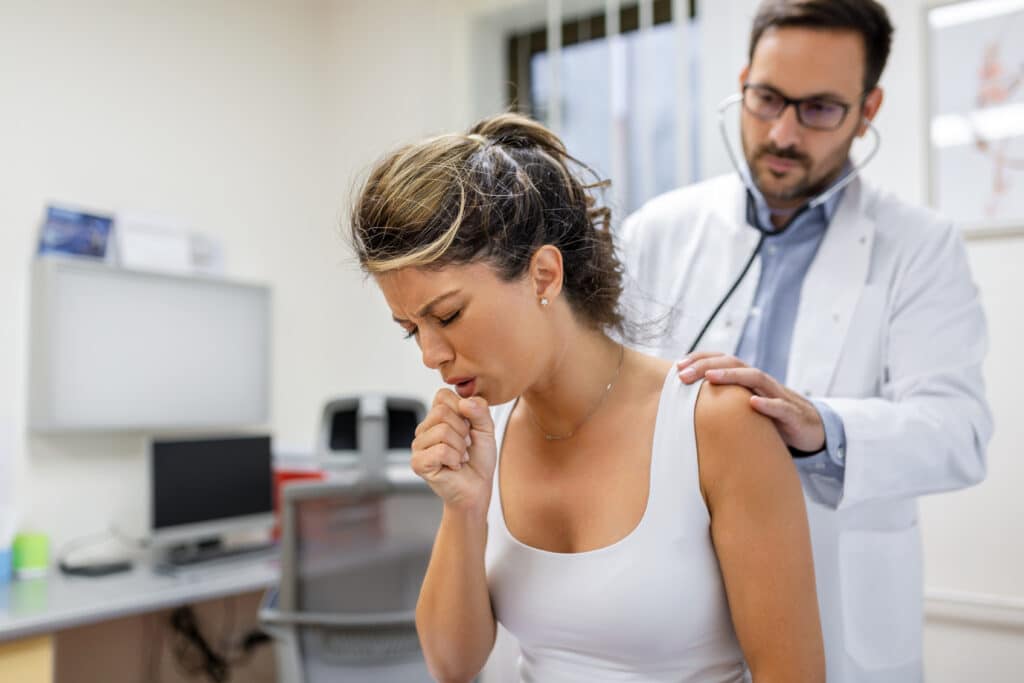 Image of a young female patient in the clinic suffering from a respiratory disease. She is coughing and the doctor is listening to the wheezing with a stethoscope.